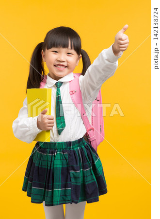 A joyful elementary school girl holding a yellow textbook and raising her thumb up in a sign of success or readiness for the new school year. 142138724