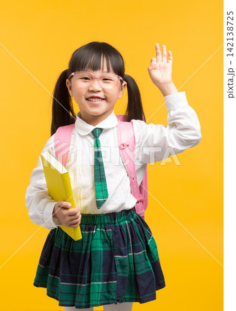 A cute schoolgirl wearing glasses waves her hand in a greeting or answering a question in class against a bright yellow background. 142138725
