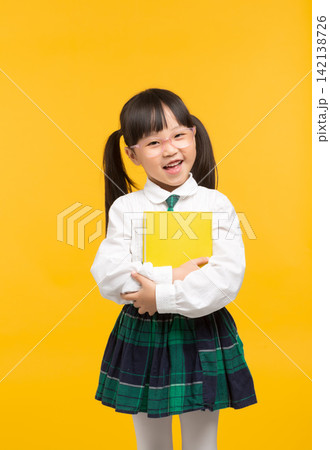 A smiling young student wearing glasses holds a yellow book tightly to her chest against a bright yellow studio background. 142138726