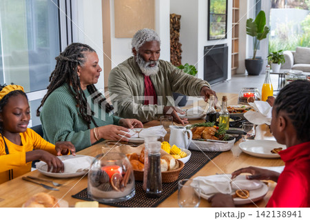 African American family passing dishes and serving roasted chicken at long wooden dining table African American family passing dishes and serving roasted chicken at long wooden dining table 142138941