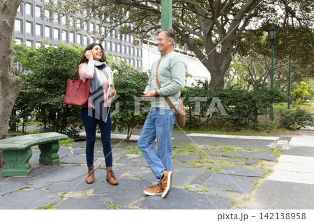 Couple standing and talking in city park holding small white flower, smartphone, red tote, satchel Couple standing and talking in city park holding small white flower, smartphone, red tote, satchel 142138958