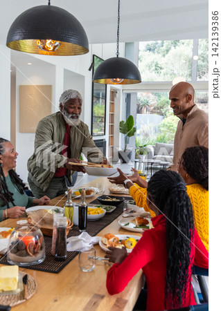 African American family with senior man passing bowl at dining table, wearing casual sweaters African American family with senior man passing bowl at dining table, wearing casual sweaters 142139386
