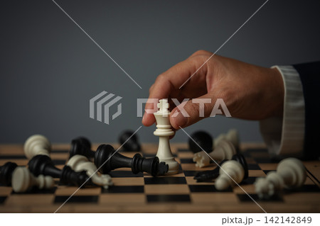 business hand holding white chess king on board with fallen pieces 142142849