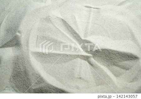Close-up of a circular geometric pattern or wheel imprint pressed into fine, light-colored sand. Summer, natural or texture background Close-up of a circular geometric pattern or wheel imprint pressed into fine, light-colored sand. Summer, natural or texture background 142143057