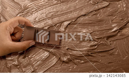 Hand holding a milk chocolate bar piece over a textured background of spread chocolate cream. World Chocolate Day celebration 142143085
