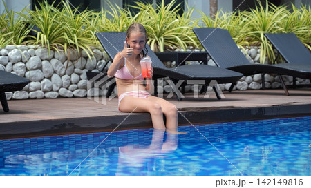 Teenage girl in pink two-piece swimsuit sitting on pool edge with legs in water giving thumbs up to camera while drinking a strawberry cocktail. Happy summer concept Teenage girl in pink two-piece swimsuit sitting on pool edge with legs in water giving thumbs up to camera while drinking a strawberry cocktail. Happy summer concept 142149816