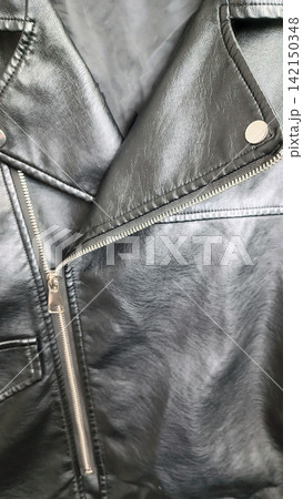 Close up of the upper left side of a black faux leather jacket, showing the lapel, diagonal silver zipper, button, seams, and stitching, with a slightly wrinkled textured surface that reflects light Close up of the upper left side of a black faux leather jacket, showing the lapel, diagonal silver zipper, button, seams, and stitching, with a slightly wrinkled textured surface that reflects light 142150348