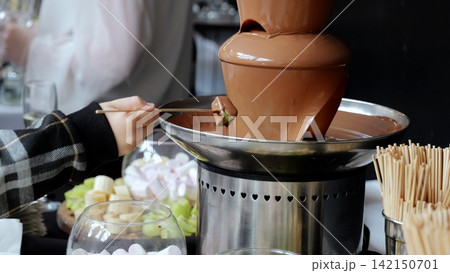 Wooden skewer piercing kiwi slice, dripping with melted chocolate, nestled among marshmallows and fruit slices during dessert celebration 142150701