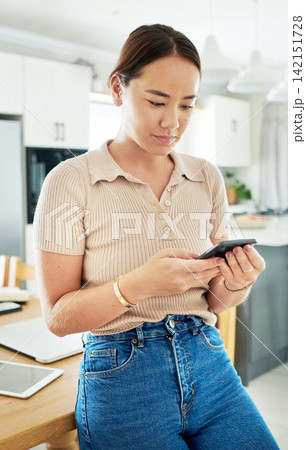Phone, reading and woman in kitchen networking on mobile app, internet or website at home. Technology, communication and young Asian female person scroll on cellphone by table at modern apartment. 142151728