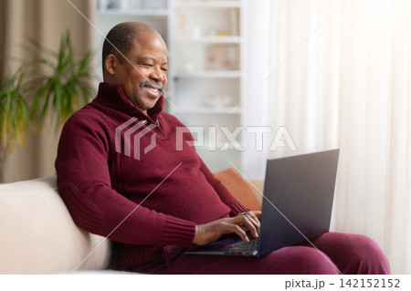Smiling mature Black man sitting on sofa at home and using laptop Smiling mature Black man sitting on sofa at home and using laptop 142152152