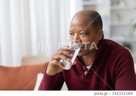 Mature Black man drinking glass of water while sitting at home 142152164