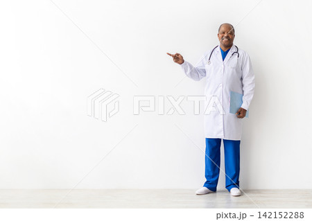 Smiling mature Black doctor in white coat pointing toward large copy space 142152288