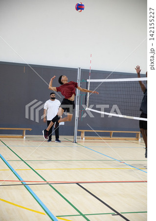 Male players, coach seeing teen player in maroon shirt jumping, spiking volleyball near net in gym Male players, coach seeing teen player in maroon shirt jumping, spiking volleyball near net in gym 142155227