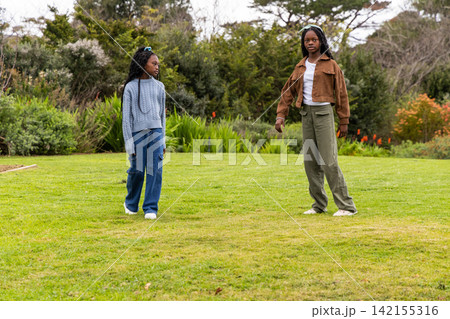 Two African American teenage girls standing on green turf in park near flowers, wearing headband 142155316