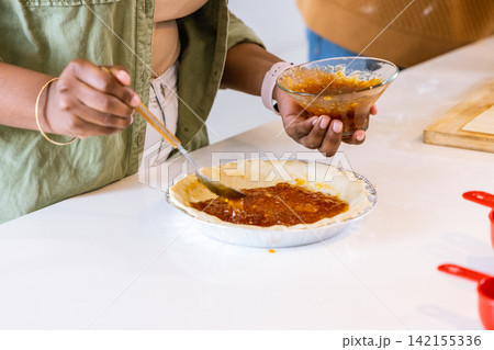 African American woman in green jacket and companion spreading jam into aluminum pie tin on counter 142155336