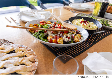 Dining table displaying oval salad bowl with two wooden spoons, lattice pie, mashed potatoes 142156010