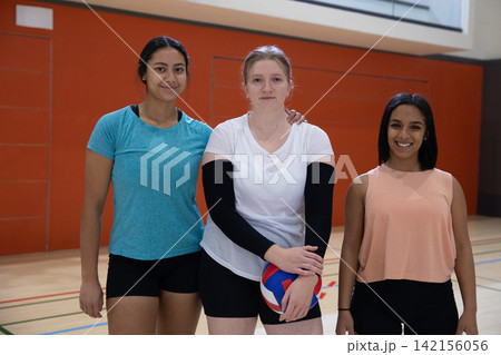 Diverse female teammates standing on gym court holding red white blue volleyball before orange wall Diverse female teammates standing on gym court holding red white blue volleyball before orange wall 142156056