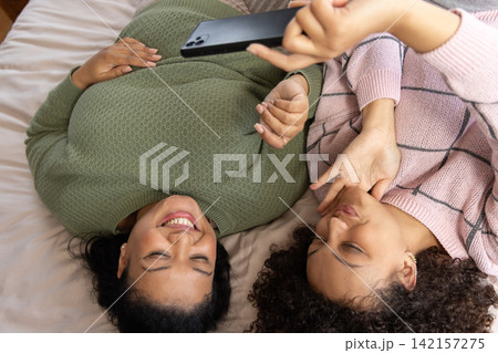 African American female friends lying on bed in knit sweaters posing for selfie, holding smartphone African American female friends lying on bed in knit sweaters posing for selfie, holding smartphone 142157275