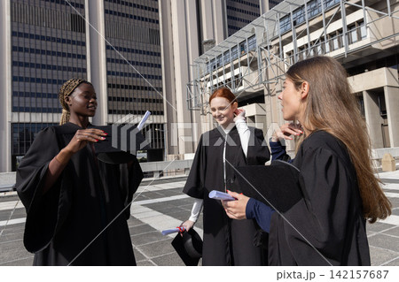 Diverse female graduates in black gowns posing on tiled plaza, holding caps and rolled diplomas 142157687