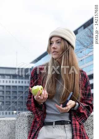 Woman wearing beanie and plaid shirt, leaning on concrete ledge in city holding apple and phone Woman wearing beanie and plaid shirt, leaning on concrete ledge in city holding apple and phone 142157688