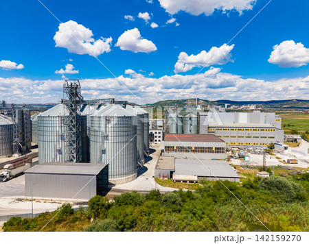 View of grain silos and industrial buildings in a rural area under a clear blue sky with white clouds 142159270
