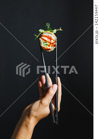Hands holding sushi with chopsticks on a black background in a food setting Hands holding sushi with chopsticks on a black background in a food setting 142159447