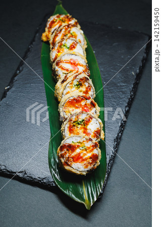 Sushi rolls arranged neatly on a dark stone background with a green leaf for decoration and a glossy sauce on top Sushi rolls arranged neatly on a dark stone background with a green leaf for decoration and a glossy sauce on top 142159450
