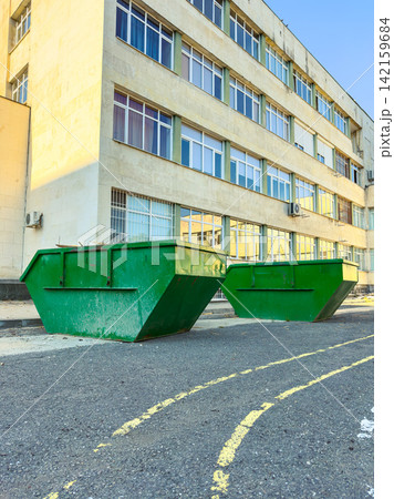 Large green waste containers sit on the asphalt surface near a building during daytime in an urban area Large green waste containers sit on the asphalt surface near a building during daytime in an urban area 142159684