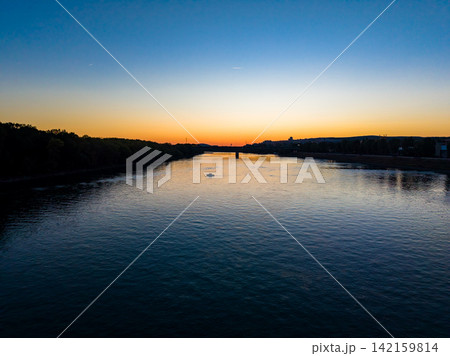River at sunset with a small boat in the water and hills in the background near a city along the riverbank River at sunset with a small boat in the water and hills in the background near a city along the riverbank 142159814