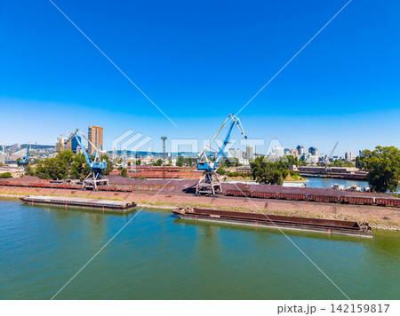 Cranes lift cargo at the river port while ships wait along the bank under a clear blue sky 142159817