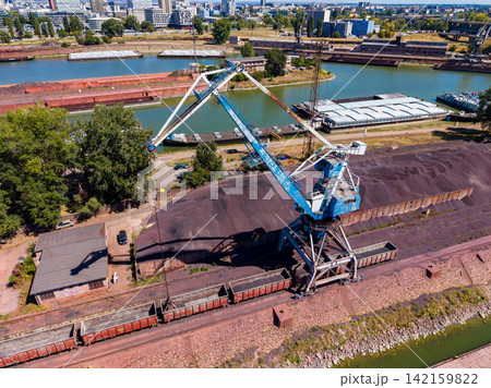 Cranes operate at a busy industrial site near water in a city on a clear day with cargo trains and stockpiled materials 142159822