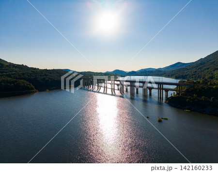 Bridge spans over lake with surrounding mountains and sun shining in the clear sky during daytime Bridge spans over lake with surrounding mountains and sun shining in the clear sky during daytime 142160233