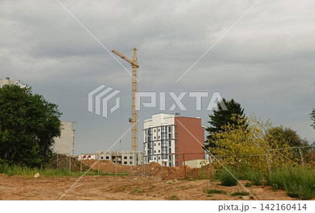 Active urban development site under a cloudy sky with red brown dirt foreground, chain link fence, and dirt piles. 142160414