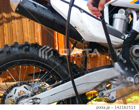 Close-up of a person bending over to hold a pressure washer gun and clean the rear wheel area of a white dirt bike outdoors. Close-up of a person bending over to hold a pressure washer gun and clean the rear wheel area of a white dirt bike outdoors. 142162513