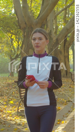 Vertical video. Sporty young woman using smartphone during breaks between runs in park. Morning Training. Healthy Lifestyle. Vertical video. Sporty young woman using smartphone during breaks between runs in park. Morning Training. Healthy Lifestyle. 142162558