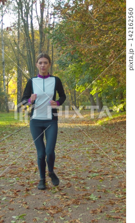 Vertical video. Active woman enjoying music while jogging in the park. Happy sportswoman enjoys her workout and stays active outdoors. 142162560