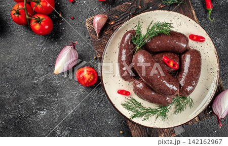 Plate of traditional blood sausages garnished with fresh herbs and red chili peppers, surrounded by ripe tomatoes and shallots on a textured dark surface. space for text 142162967