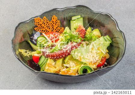 Colorful salad with octopus, fresh vegetables, and decorative elements served in a black bowl on a gray surface Colorful salad with octopus, fresh vegetables, and decorative elements served in a black bowl on a gray surface 142163011