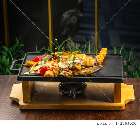 Sizzling seafood platter with shrimp, scallops, and vegetables served on a black iron grill atop a wooden board, garnished with herbs and spices 142163020