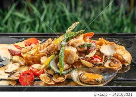 Grilled seafood platter with shrimp, mussels, and vegetables served on a sizzling plate, garnished with green asparagus and cherry tomatoes against a blurred green background Grilled seafood platter with shrimp, mussels, and vegetables served on a sizzling plate, garnished with green asparagus and cherry tomatoes against a blurred green background 142163021