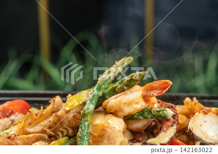 Grilled seafood dish featuring shrimp, octopus, and asparagus garnished with colorful vegetables served on a black plate with a blurred green background 142163022