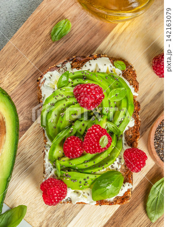 Sliced whole grain bread topped with creamy cheese, avocado, fresh raspberries, and basil leaves on a wooden cutting board with olive oil in the background 142163079