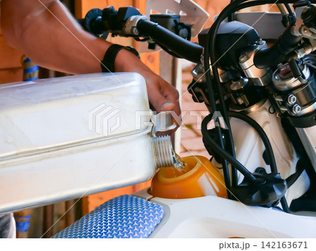 Man pouring fuel into a motorcycle tank from a white canister. Maintenance and refueling of a motorbike in a garage. Preparation for a ride or adventure. Automotive mechanical service. 142163671