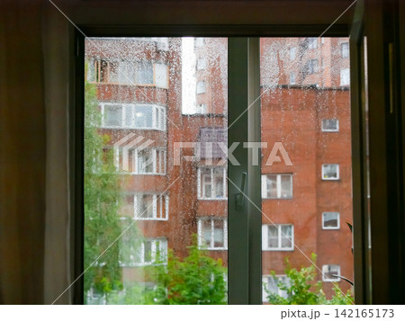 Raindrops on window glass looking out at city apartment building. Gloomy weather outside home view. Melancholy concept of staying indoor during a storm with autumn atmosphere. Raindrops on window glass looking out at city apartment building. Gloomy weather outside home view. Melancholy concept of staying indoor during a storm with autumn atmosphere. 142165173