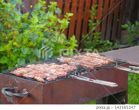 Close-up of meat cooking on a rustic rusty metal barbecue grill outdoors in a garden, with visible smoke and steam. 142165347