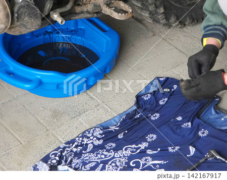 Man mechanic wearing gloves changing oil in vehicle. Automotive maintenance service with blue drain pan. Professional repairman working on dirty machine motor in garage workshop environment. Man mechanic wearing gloves changing oil in vehicle. Automotive maintenance service with blue drain pan. Professional repairman working on dirty machine motor in garage workshop environment. 142167917