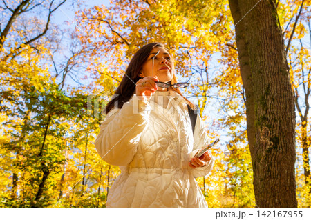 Woman walking in autumn park with mobile phone. Girl enjoying sunny fall foliage in woods. Relaxed lady looking away while holding smartphone. Lifestyle outdoor leisure activity concept. Woman walking in autumn park with mobile phone. Girl enjoying sunny fall foliage in woods. Relaxed lady looking away while holding smartphone. Lifestyle outdoor leisure activity concept. 142167955