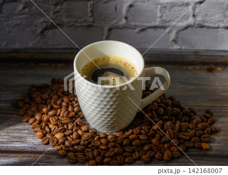 Warm cup of coffee resting on fresh coffee beans with rustic wooden backdrop 142168007