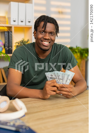 African American young man on home table counting cash money smiling after profit or winning deal African American young man on home table counting cash money smiling after profit or winning deal 142170090