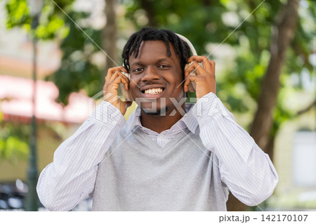 African American young man putting on wireless headphones walking in city street smiling at camera African American young man putting on wireless headphones walking in city street smiling at camera 142170107